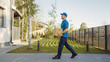 © Gorodenkoff - Delivery Man Holding Card Board Package Enters Through the Gates and Walks to the House and Knocks. Delivering Postal Parcel. In the Background Beautiful Suburban Neighbourhood. Side View