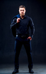 Wall Mural - Fashion portrait of young man in black shirt. Man poses over dark wall with one hand in pocket. Handsome confident man, businessman, modern clothes.