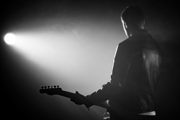 Rock guitarist man in leather jacket standing his back in smoky studio or stage masterfully playing electric guitar. View of unrecognizable musician in the spotlight.