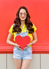 Wall Mural - Beautiful woman holding a red heart over red background. Fashion portrait stylish pretty woman outdoor.