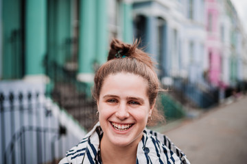 Portrait of an attractive natural young girl smiling with green eyes, blonde hair and blue and white striped jacket, on a street with colorful houses in London.