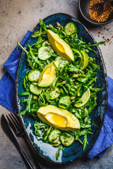 Wall Mural - Green salad with cucumber, avocado and arugula in blue dish, dark background. top view.