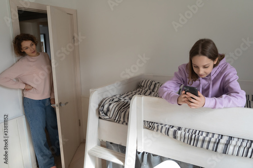 Worried adult parent mom coming to teenage child bedroom unhappy about technology addicted kid using phone, obsessed with mobile social media apps. Teens gadgets overuse and parental control concept.