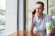 © DragonImages - Handosme young man sitting at cafe table, drinking coffe and talking on phone with colleague