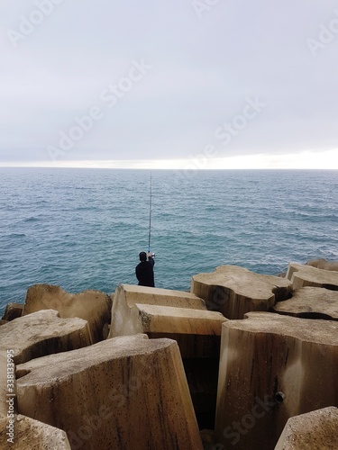 Fotografía Rear View Of Mature Man Fishing In Sea While Sitting On Rock Against Cloudy Sky