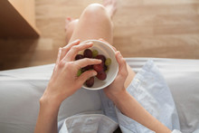 A young woman holding a bowl of grapes on her legs in her room at home and sunlight coming through the window