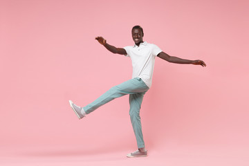 Smiling young african american man guy in white polo shirt, turquoise trousers isolated on pastel pink background. People lifestyle concept. Mock up copy space. Spreading hands, rising leg, dancing.