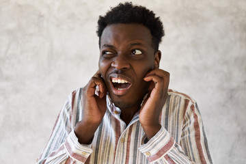 Wall Mural - This is too loud! Studio image of stressed frustrated young dark skinned male grimacing and plugging ears with fingers, can't stand noise, having painful facial expression, trying to fall asleep