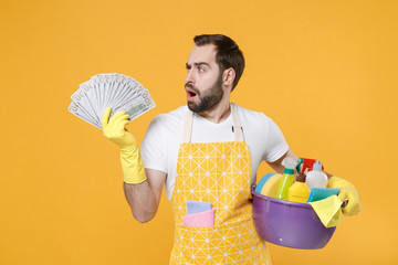 Puzzled young man househusband in apron rubber gloves hold basin with detergent bottles washing cleansers doing housework isolated on yellow background. Housekeeping concept. Hold fan of cash money.