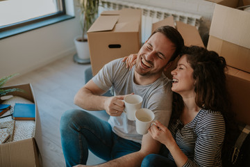 Top view photo of young happy couple drinking coffee while cardboard boxes are all around them. Young couple is moving in to their new home.