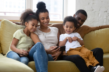 Wall Mural - African american smiling family with two children having good time using smartphone. Young happy diverse husband and wife with cute daughter and son using applications in mobile phone.