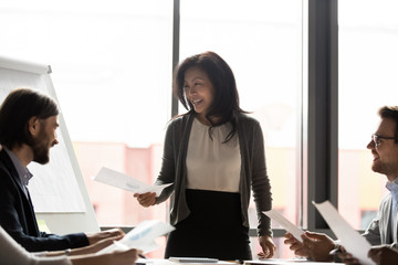 Smiling middle-aged Asian team leader female boss stand lead office meeting with diverse colleagues share financial documents handouts, happy ethnic businesswoman head group briefing with coworkers