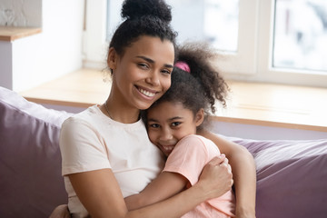 Wall Mural - Close up headshot portrait of millennial african american mother and teen daughter looking at camera sitting on couch. Happy smiling diverse family embrace together in living room at home.