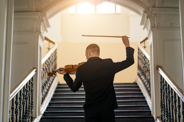 rear view on young caucasian male in elegant stylish suit playing violin in opposite of stairs. professional classical music performer