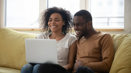 Wall Mural - Happy african american family having good time using laptop. Young smiling husband and wife sitting on couch at home looking at computer screen and watching video.