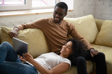 Young happy african american couple watching at tablet screen. Attractive millennial family of diverse woman lies on man sitting on sofa. Husband and wife using mobile device for online shopping.