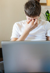 Cute upset boy in white t shirt sitting on the couch in the living room next to laptop and study. Stressed. tired kid. Home education, self education by kids.	