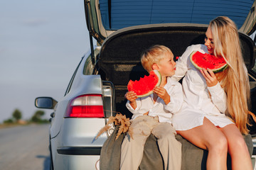 pretty blond hair woman with little blond son at sunset relaxing behind the car and eating watermelon. summer, travel, nature and fresh air in the countryside.