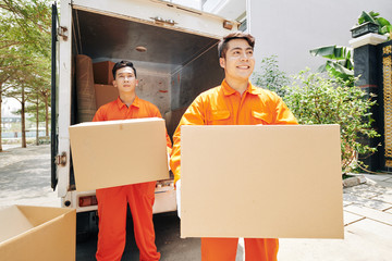 Wall Mural - Positive Vietnamese moving service workers in orange uniform carrying cardboard boxes to house entrance