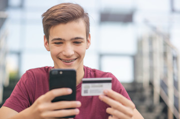 teenage boy with a credit card and mobile phone makes purchasing outdoors. happy young man is using 