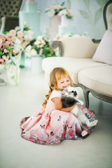 Vertical picture of cute smiling blonde 3 years old girl in pink dress sitting on floor with funny furry gray and white rabbit. Small bunny and happy child. Easter rabbit and small kid. Indoor