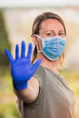 Stop COVID-19. Outdoor portrait of a blonde woman with protective medical mask and gloves shows the stop coronavirus sign with her hand, looking at the camera. Pandemic concept.