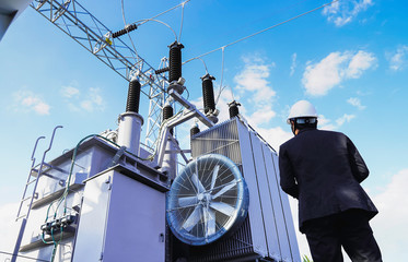 A low angle image of a businessman wearing a black suit, standing looking at a large power transformer with blue sky to be background, Concept about business people who want to invest in energy