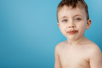 Wall Mural - Little boy eating donut chocolate on blue background. Cute happy boy smeared with chocolate around his mouth. Child concept, tasty food for kids