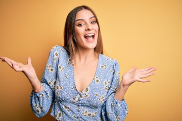 Young beautiful brunette woman wearing casual floral t-shirt standing over yellow background celebrating crazy and amazed for success with arms raised and open eyes screaming excited. Winner concept