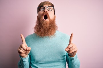 Poster - Handsome Irish redhead man with beard wearing glasses over pink isolated background amazed and surprised looking up and pointing with fingers and raised arms.