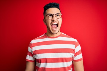 Young handsome man wearing casual striped t-shirt and glasses over isolated red background angry and mad screaming frustrated and furious, shouting with anger. Rage and aggressive concept.