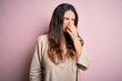 Young beautiful brunette woman wearing casual sweater standing over pink background smelling something stinky and disgusting, intolerable smell, holding breath with fingers on nose. Bad smell
