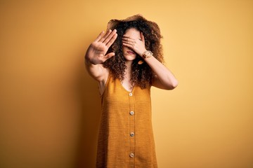 Beautiful brunette woman on vacation with curly hair and piercing wearing hat and dress covering eyes with hands and doing stop gesture with sad and fear expression. Embarrassed and negative concept.