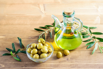 Wall Mural - Olive oil in the glass bottle, olives in the bowl and branches of olive tree on the wooden background in the sunlights.