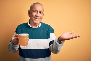 Sticker - Senior handsome man drinking jar of beer standing over isolated yellow background smiling cheerful presenting and pointing with palm of hand looking at the camera.