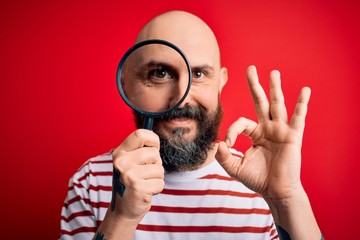 Poster - Handsome detective bald man with beard using magnifying glass over red background doing ok sign with fingers, excellent symbol