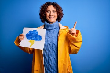 Poster - Middle age curly hair woman wearing rain coat holding banner with cloud and thunder surprised with an idea or question pointing finger with happy face, number one