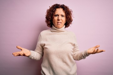 Poster - Middle age beautiful curly hair woman wearing casual turtleneck sweater over pink background clueless and confused with open arms, no idea concept.