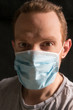 Close up selfie of a redheaded man wearing a surgical respiratory mask against COVID-19 coronavirus, background is dark.