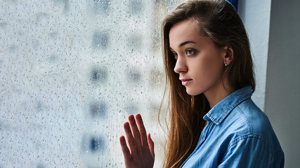 Wall Mural - Portrait of a young attractive brunette lonely sad melancholy pensive caucasian woman with long hair standing alone near the window with raindrops in rainy autumn weather