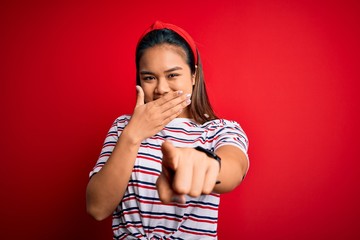 Young beautiful asian girl wearing casual striped t-shirt over isolated red background laughing at you, pointing finger to the camera with hand over mouth, shame expression