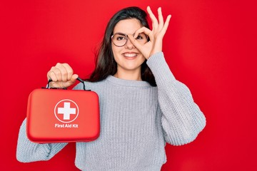 Young beautiful girl holding first aid kit medical box over red background with happy face smiling doing ok sign with hand on eye looking through fingers