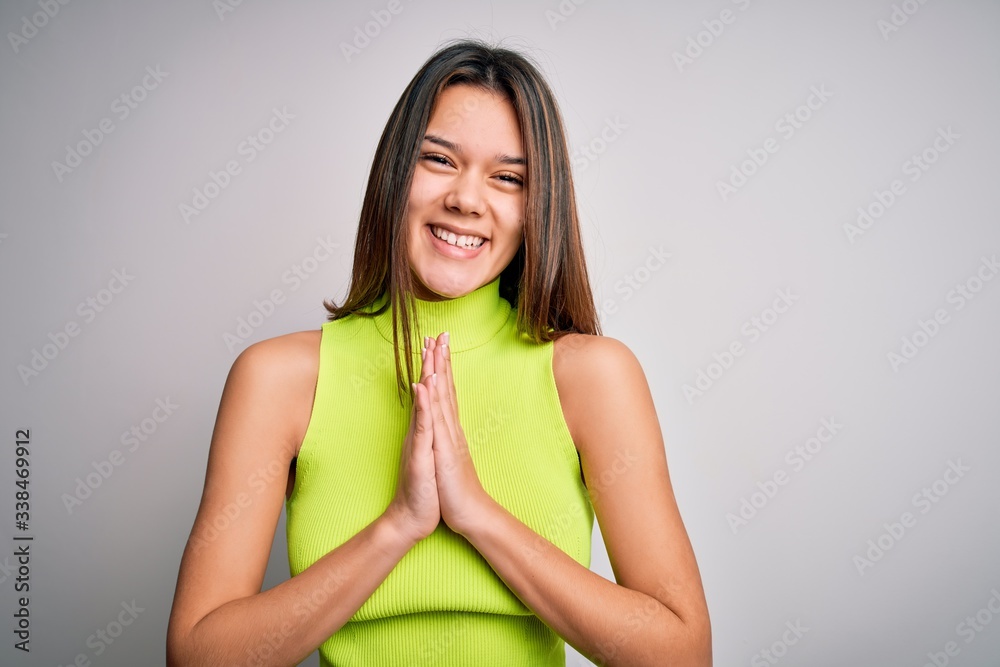 Young beautiful brunette girl wearing casual summer t-shirt over isolated white background praying with hands together asking for forgiveness smiling confident.