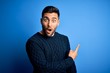 © Krakenimages.com - Young handsome man wearing casual sweater standing over isolated blue background Surprised pointing with finger to the side, open mouth amazed expression.