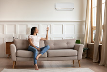 Young Asian Woman Switching On Air Conditioner While Resting Seated On Couch In Modern Interior Living Room, Holds Remote Climate Control Cooler System Manage Temperature At Contemporary Home Concept