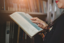 Close Up Of A Woman Hands Holding And Read The Holy Bible Overs Blurred Books In The Bookshelf, Selective Focus On Woman Hand's 