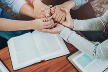 Close Up Of  People Group Holding A Hand And Pray Together Over A Blurred Holy Bible On Wooden Table, Christian Fellowship  Or Praying Meeting In Home Concept With Copy Space