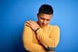 © Krakenimages.com - Young handsome latin man wearing yellow casual sweater over isolated blue background Hugging oneself happy and positive, smiling confident. Self love and self care