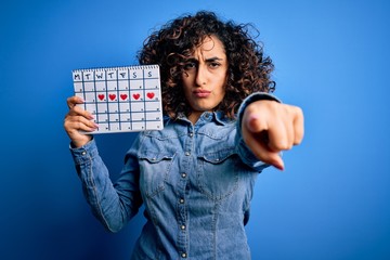 Sticker - Young beautiful curly arab woman holding period calendar controlling menstrual cycle pointing with finger to the camera and to you, hand sign, positive and confident gesture from the front