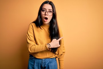 Young brunette woman wearing glasses and casual sweater over yellow isolated background Surprised pointing with finger to the side, open mouth amazed expression.